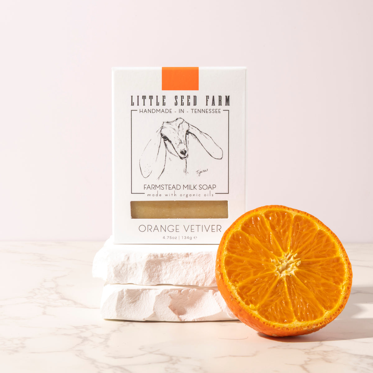 A bar of Little Seed Farm Orange Vetiver goat's milk soap sitting on stones with an orange slice.