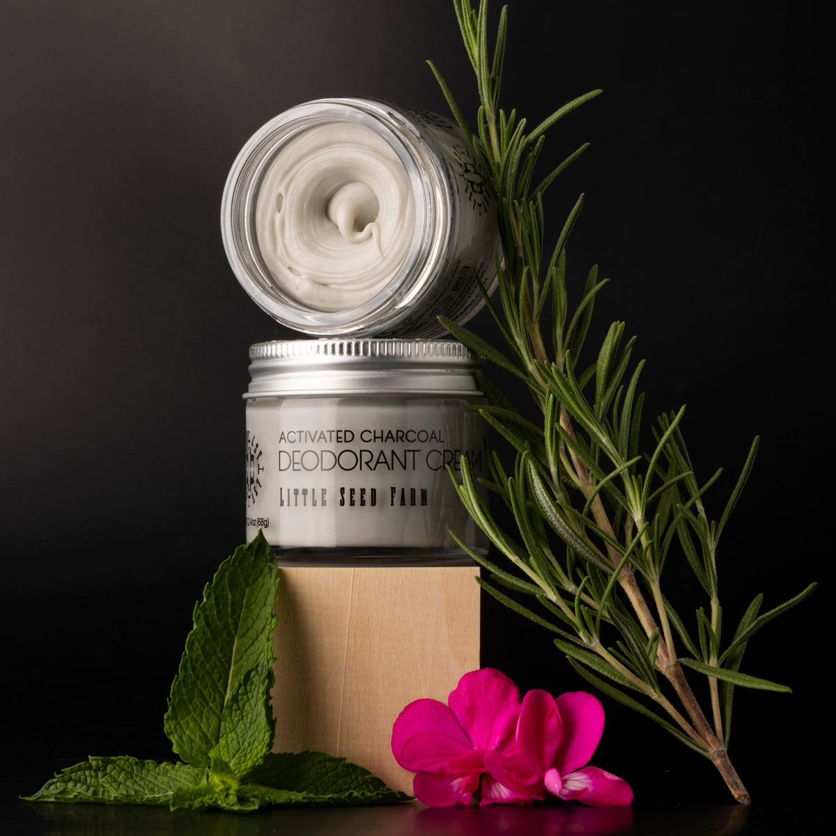 A jar of little seed farm activated charcoal deodorant cream on black background surrounded by rosemary, mint and geraniums.